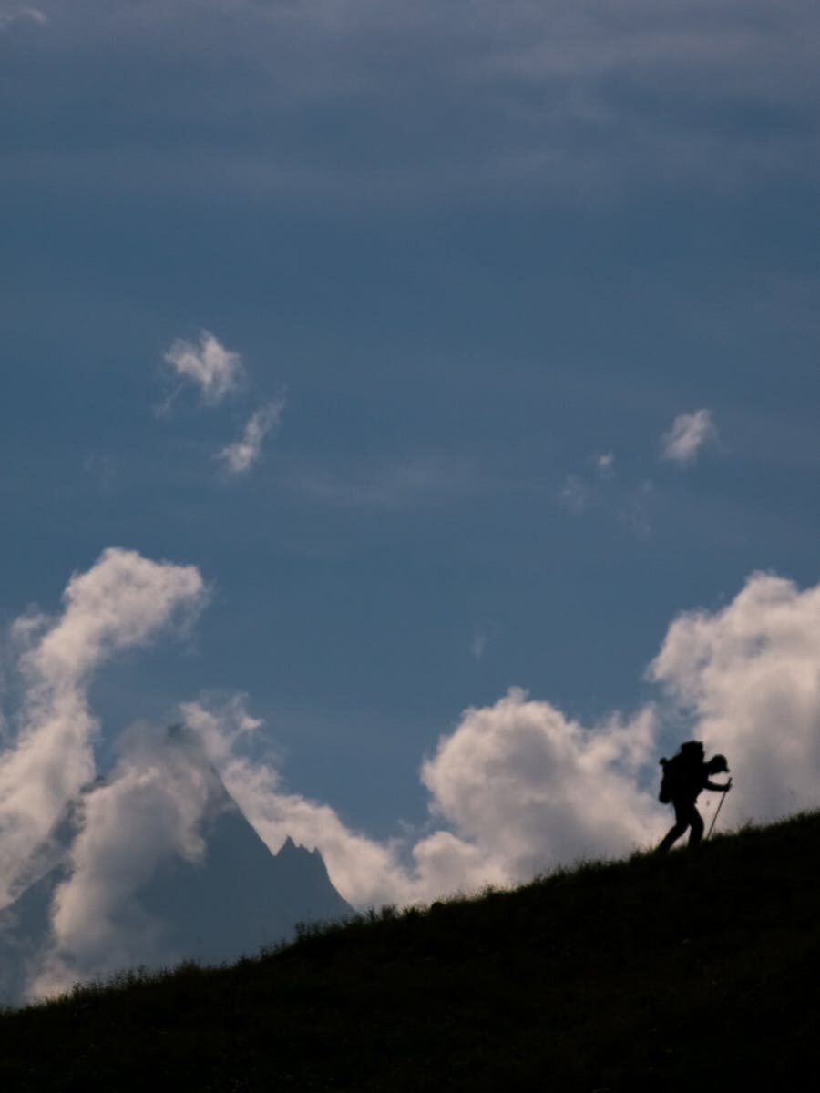 Silhouette of a hiker with trekking poles hiking up a hill against a cloudy sky in Kishtwar.